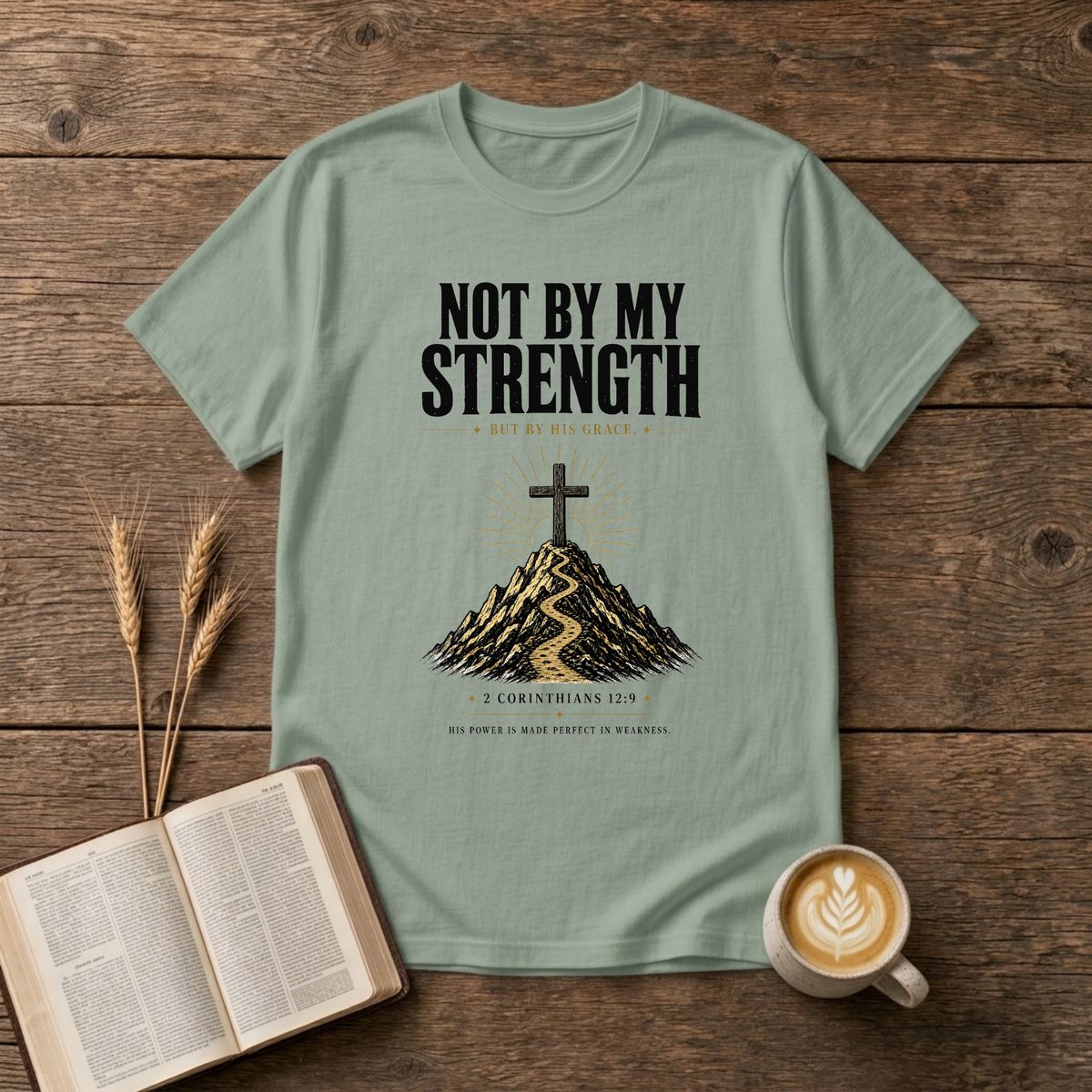 Distressed NOT BY MY STRENGTH lettering above a cross and mountain motif on a Sage unisex T-Shirt