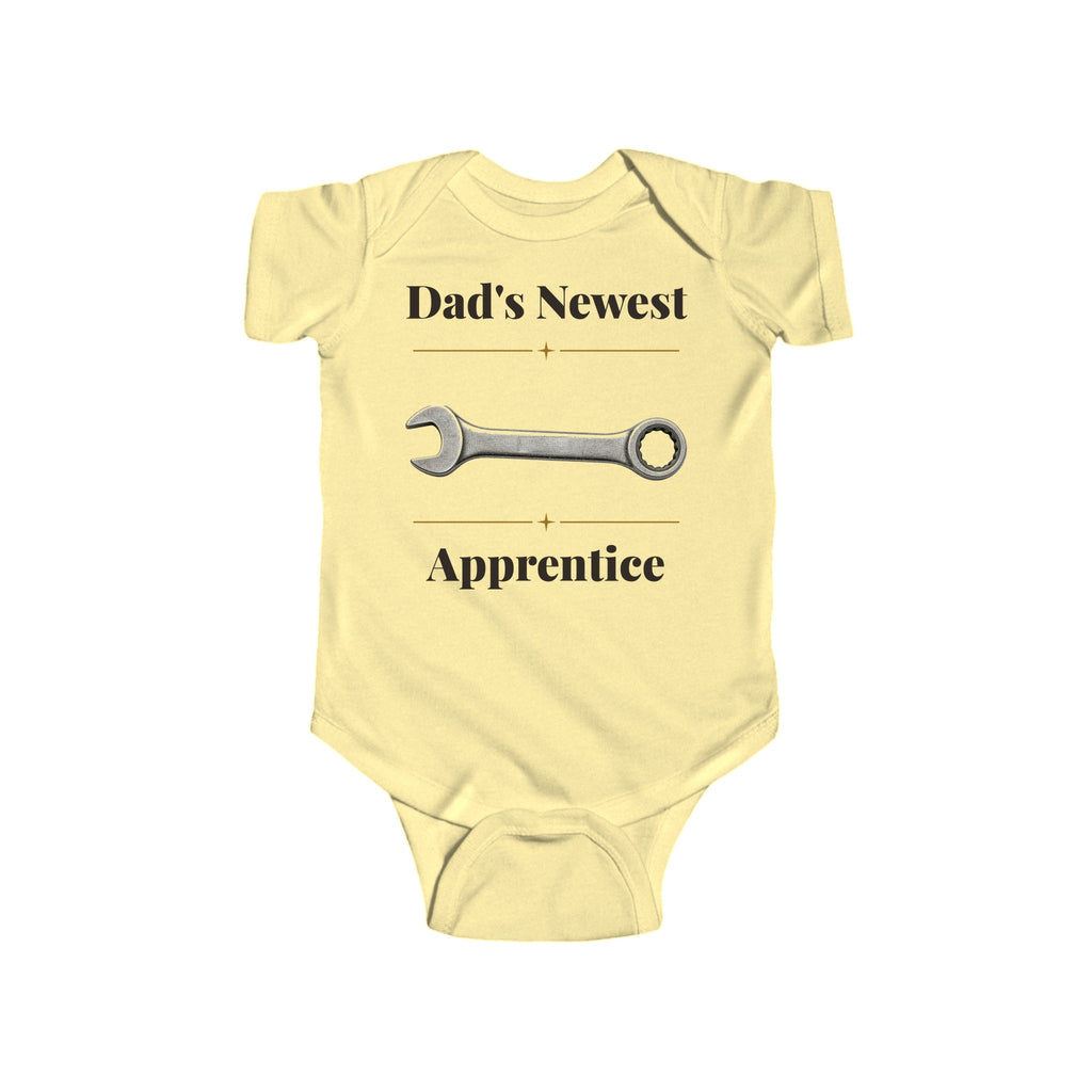 Dad's Newest Apprentice baby bodysuit in Butter on a wood table with a knit blanket, wooden rattle and open Bible