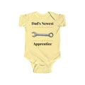 Dad's Newest Apprentice baby bodysuit in Butter on a wood table with a knit blanket, wooden rattle and open Bible