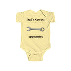 Dad's Newest Apprentice baby bodysuit in Butter on a wood table with a knit blanket, wooden rattle and open Bible