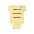 Kitchen Crew in Training baby bodysuit in Butter on a wood table with a knit blanket, wooden rattle and open Bible