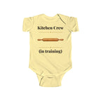 Kitchen Crew in Training baby bodysuit in Butter on a wood table with a knit blanket, wooden rattle and open Bible