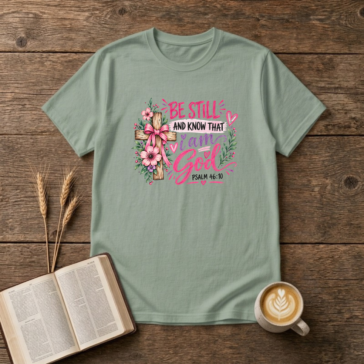 Be Still and Know That I Am God Psalm 46:10 design with a wooden cross wrapped in a pink bow and pink and purple flowers on a Sage unisex T-Shirt