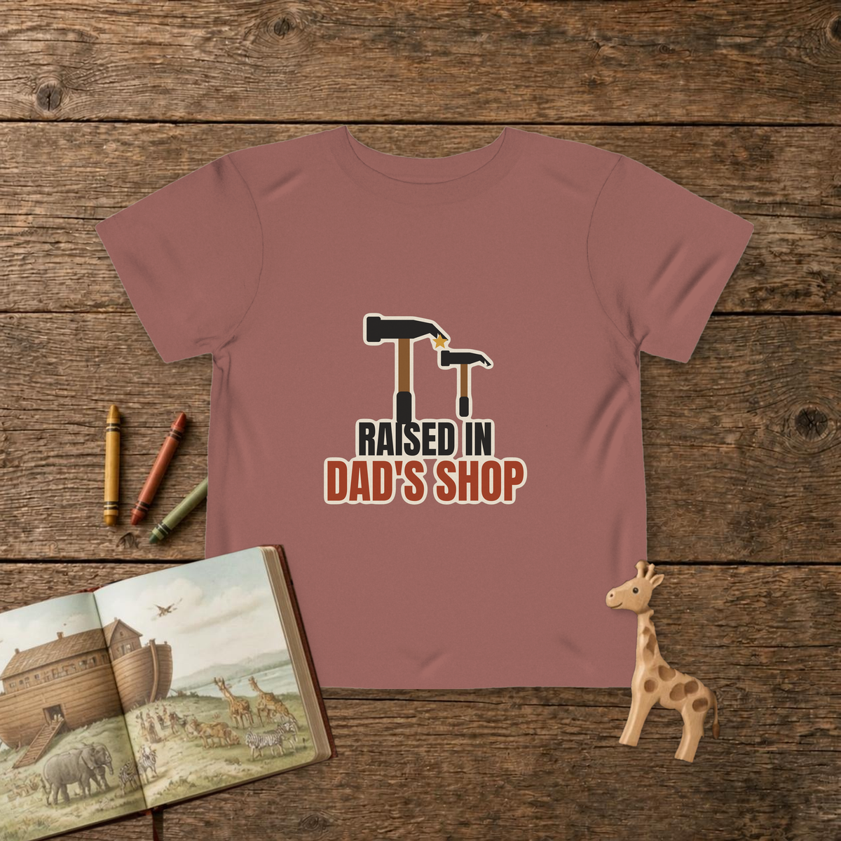 Raised In Dad's Shop toddler tee in Heather Mauve, big claw hammer next to a little hammer with a gold star, on a wooden table with a picture Bible, wooden giraffe and crayons