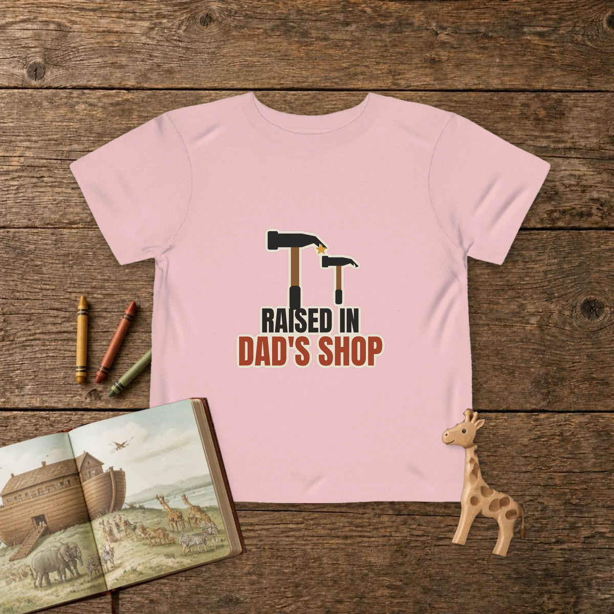 Raised In Dad's Shop toddler tee in Pink, big claw hammer next to a little hammer with a gold star, on a wooden table with a picture Bible, wooden giraffe and crayons