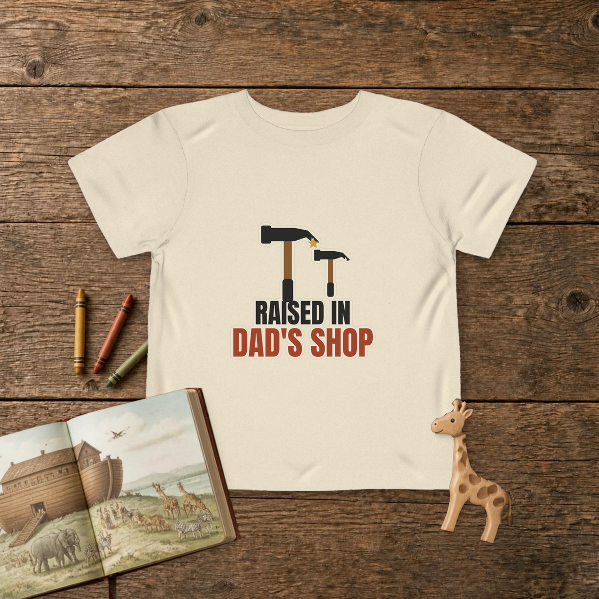 Raised In Dad's Shop toddler tee in Natural, big claw hammer next to a little hammer with a gold star, on a wooden table with a picture Bible, wooden giraffe and crayons
