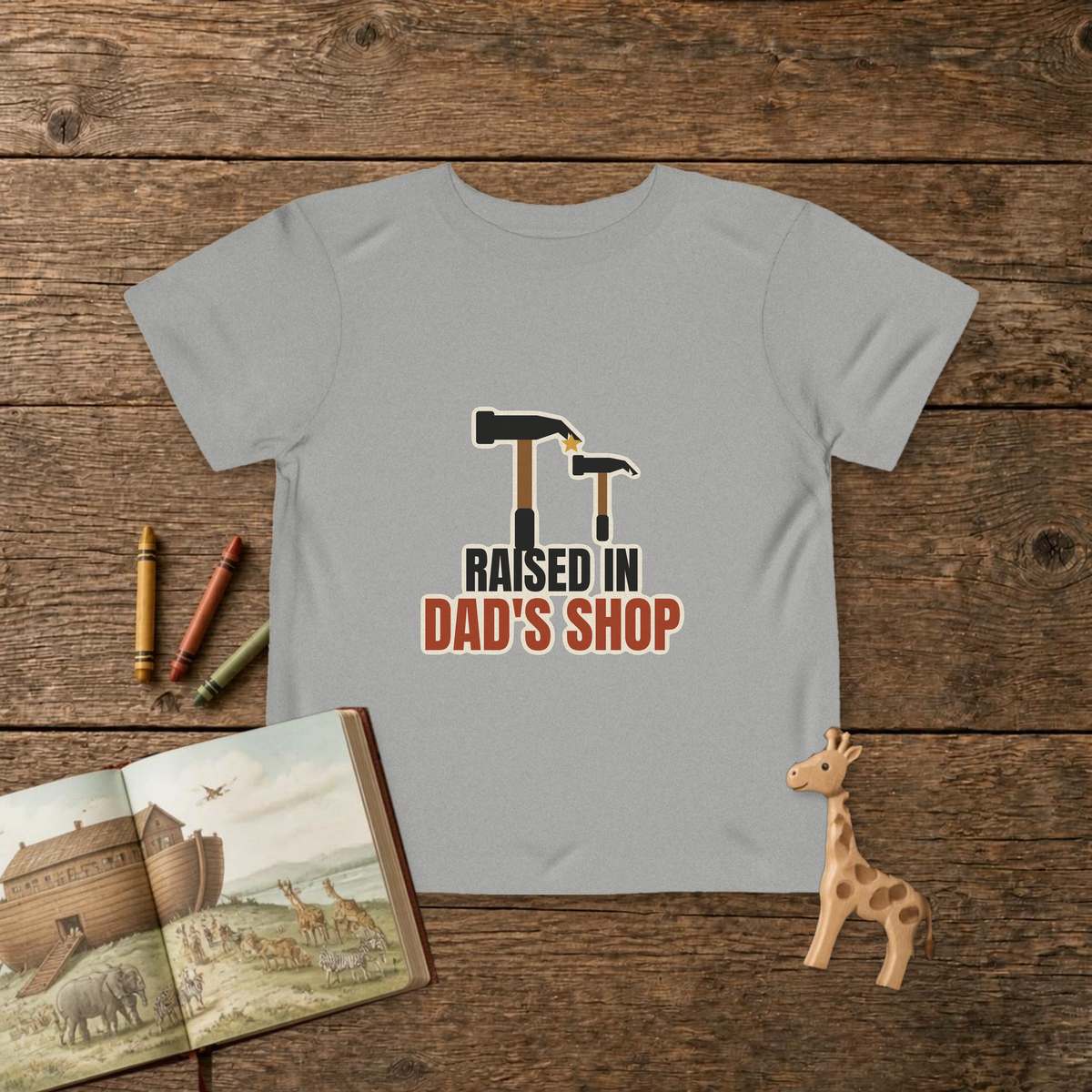 Raised In Dad's Shop toddler tee in Athletic Heather, big claw hammer next to a little hammer with a gold star, on a wooden table with a picture Bible, wooden giraffe and crayons