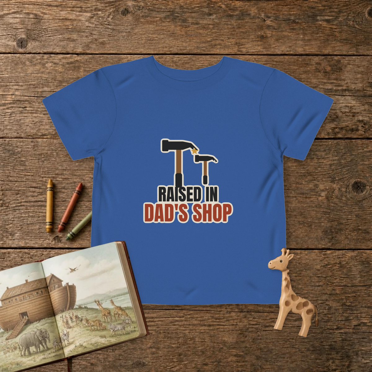 Raised In Dad's Shop toddler tee in True Royal, big claw hammer next to a little hammer with a gold star, on a wooden table with a picture Bible, wooden giraffe and crayons
