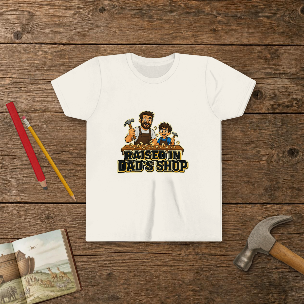 Raised In Dad's Shop youth tee in White (Cartoon design), cartoon of a dad and his young helper at a workbench with hammers in hand, with wood curls and bold "Raised In Dad's Shop" lettering, on a wooden table with a picture Bible, a child's hammer, and two pencils