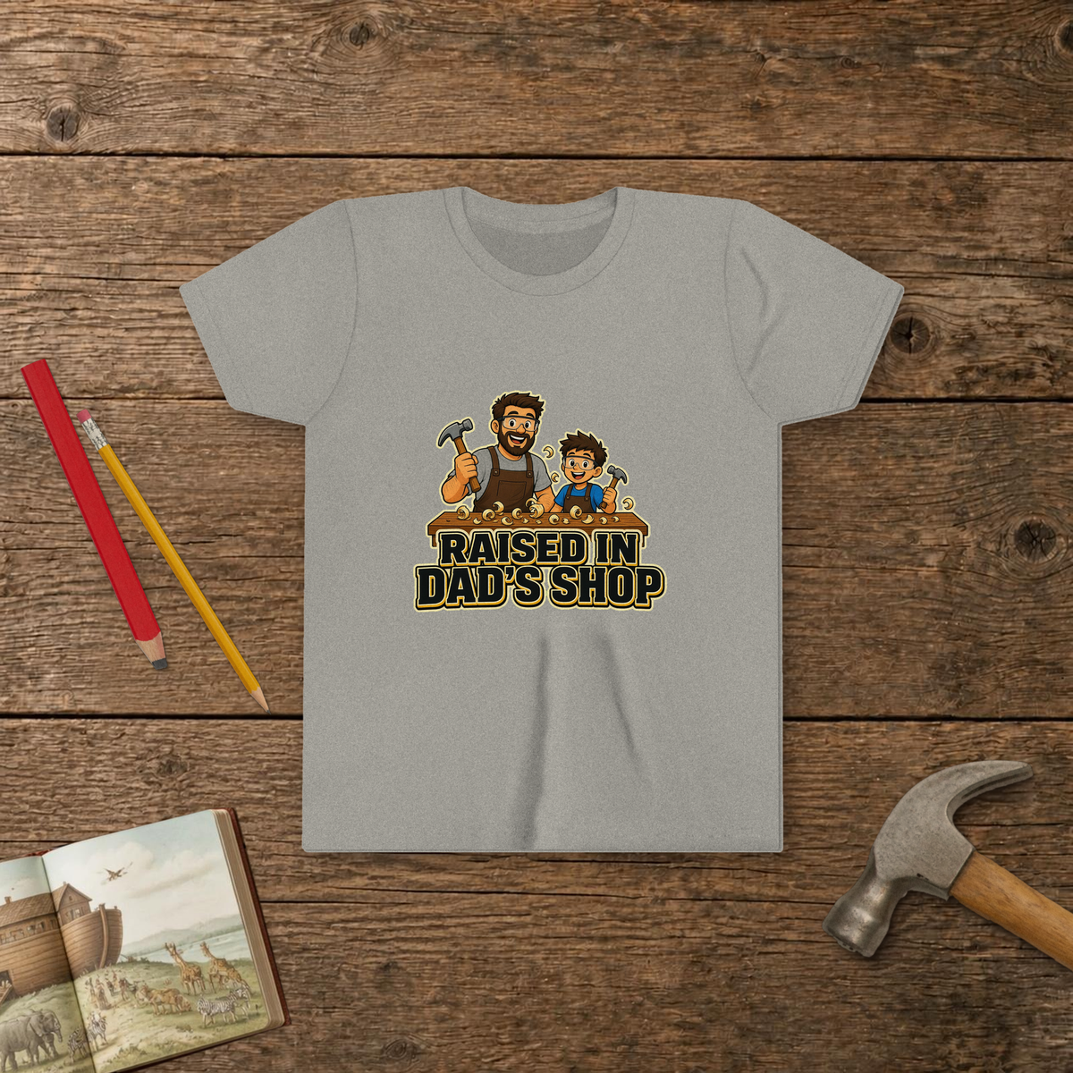 Raised In Dad's Shop youth tee in Athletic Heather (Cartoon design), cartoon of a dad and his young helper at a workbench with hammers in hand, with wood curls and bold "Raised In Dad's Shop" lettering, on a wooden table with a picture Bible, a child's hammer, and two pencils