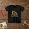 Raised In Dad's Shop youth tee in Black (Cartoon design), cartoon of a dad and his young helper at a workbench with hammers in hand, with wood curls and bold "Raised In Dad's Shop" lettering, on a wooden table with a picture Bible, a child's hammer, and two pencils