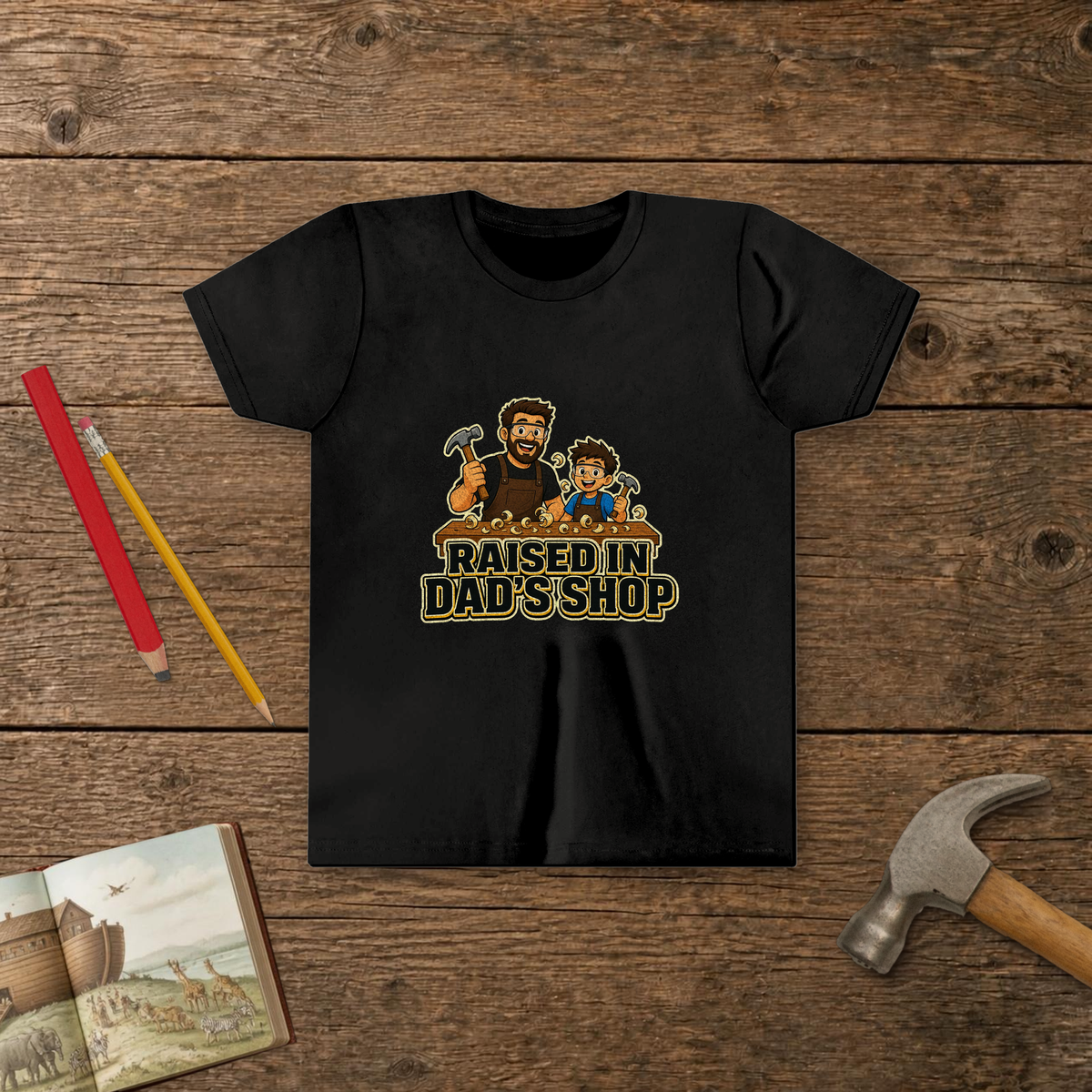 Raised In Dad's Shop youth tee in Black (Cartoon design), cartoon of a dad and his young helper at a workbench with hammers in hand, with wood curls and bold "Raised In Dad's Shop" lettering, on a wooden table with a picture Bible, a child's hammer, and two pencils