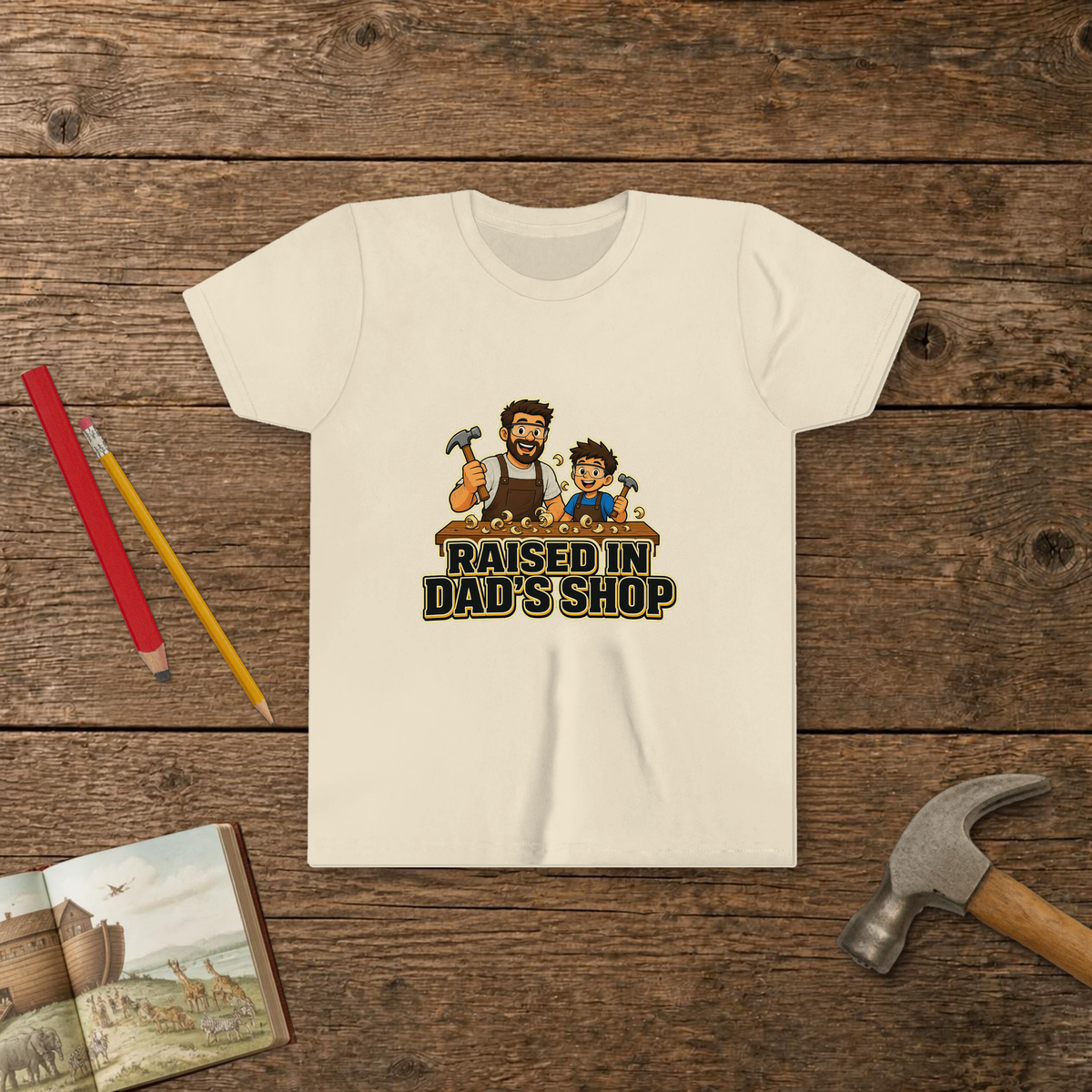 Raised In Dad's Shop youth tee in Natural (Cartoon design), cartoon of a dad and his young helper at a workbench with hammers in hand, with wood curls and bold "Raised In Dad's Shop" lettering, on a wooden table with a picture Bible, a child's hammer, and two pencils
