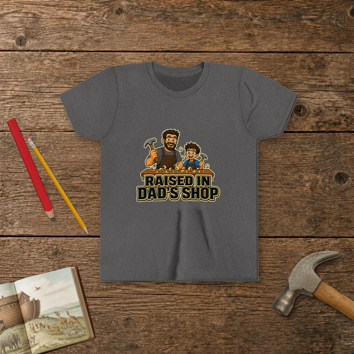 Raised In Dad's Shop youth tee in Deep Heather (Cartoon design), cartoon of a dad and his young helper at a workbench with hammers in hand, with wood curls and bold "Raised In Dad's Shop" lettering, on a wooden table with a picture Bible, a child's hammer, and two pencils
