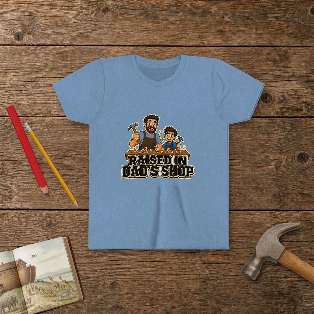 Raised In Dad's Shop youth tee in Heather Columbia Blue (Cartoon design), cartoon of a dad and his young helper at a workbench with hammers in hand, with wood curls and bold "Raised In Dad's Shop" lettering, on a wooden table with a picture Bible, a child's hammer, and two pencils