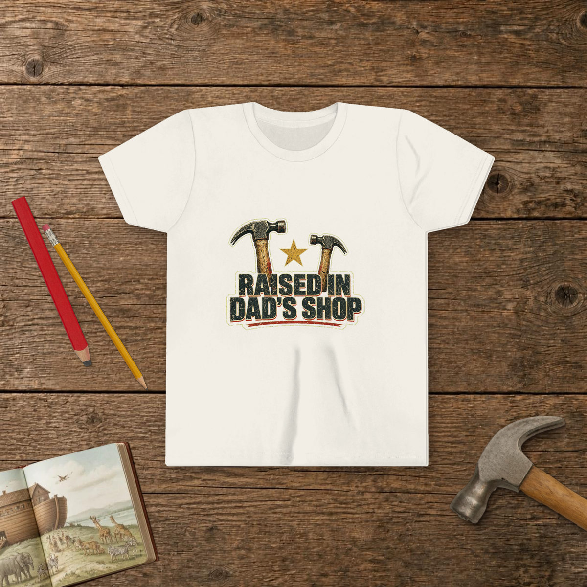 Raised In Dad's Shop youth tee in White (Vintage Hammers design), two worn claw hammers around a gold star above bold distressed "Raised In Dad's Shop" lettering in a vintage print look, on a wooden table with a picture Bible, a child's hammer, and two pencils