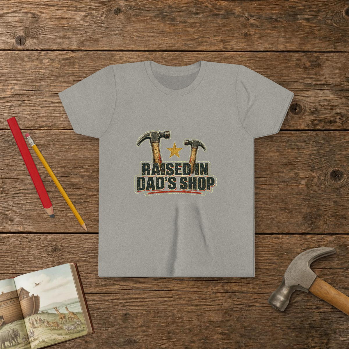 Raised In Dad's Shop youth tee in Athletic Heather (Vintage Hammers design), two worn claw hammers around a gold star above bold distressed "Raised In Dad's Shop" lettering in a vintage print look, on a wooden table with a picture Bible, a child's hammer, and two pencils