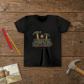 Raised In Dad's Shop youth tee in Black (Vintage Hammers design), two worn claw hammers around a gold star above bold distressed "Raised In Dad's Shop" lettering in a vintage print look, on a wooden table with a picture Bible, a child's hammer, and two pencils