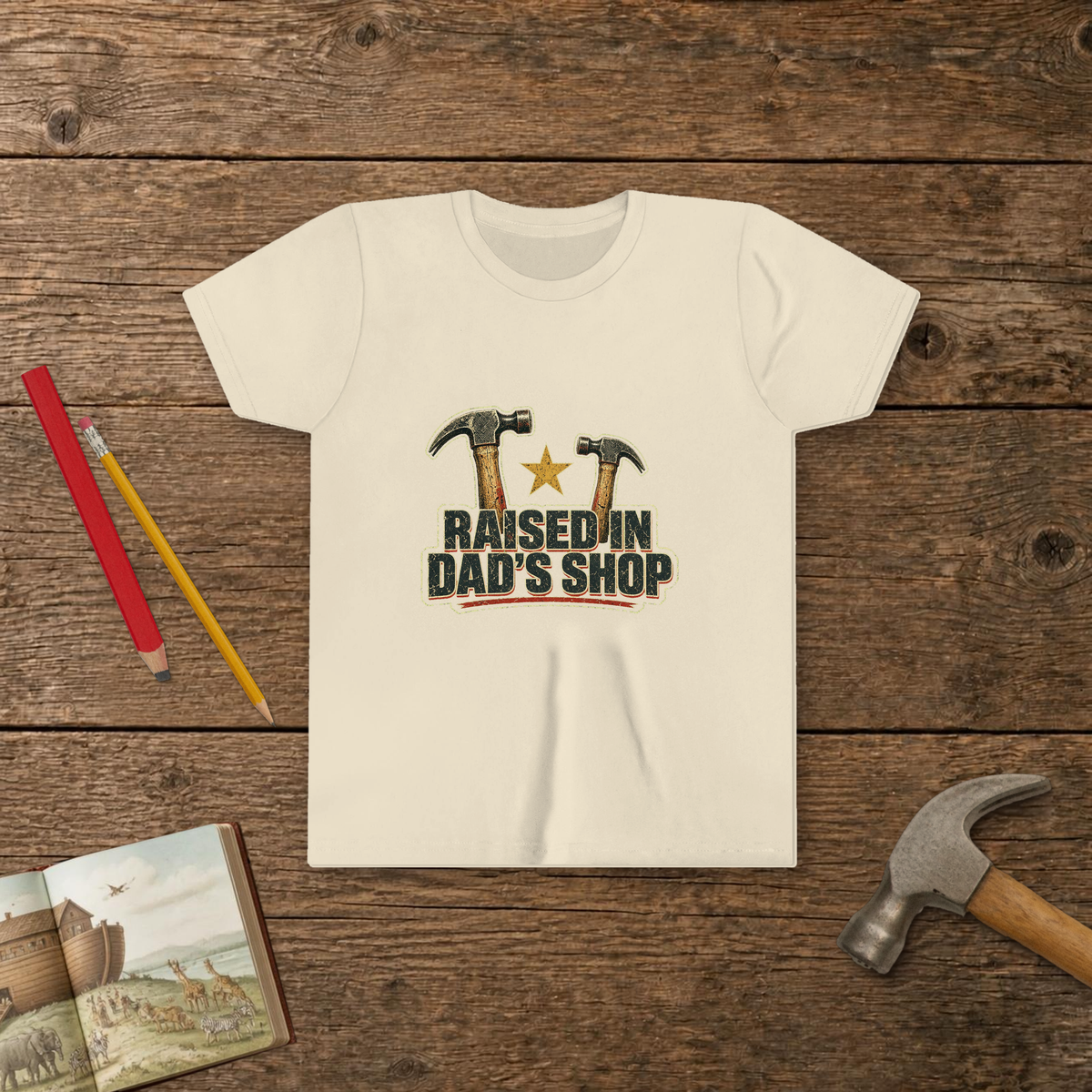 Raised In Dad's Shop youth tee in Natural (Vintage Hammers design), two worn claw hammers around a gold star above bold distressed "Raised In Dad's Shop" lettering in a vintage print look, on a wooden table with a picture Bible, a child's hammer, and two pencils