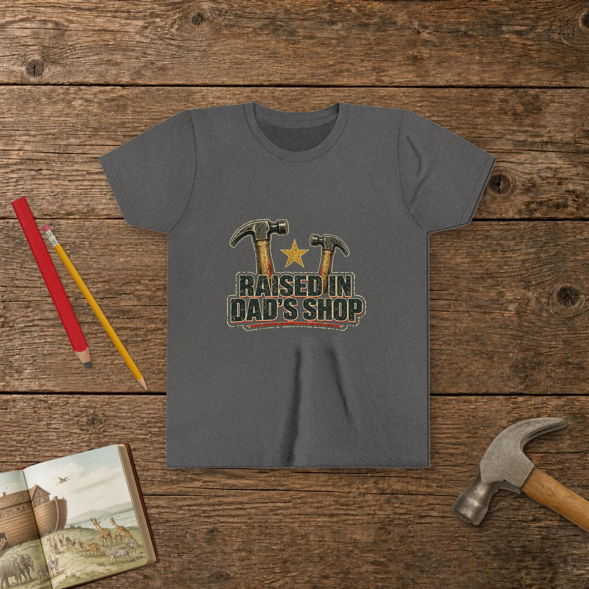 Raised In Dad's Shop youth tee in Deep Heather (Vintage Hammers design), two worn claw hammers around a gold star above bold distressed "Raised In Dad's Shop" lettering in a vintage print look, on a wooden table with a picture Bible, a child's hammer, and two pencils