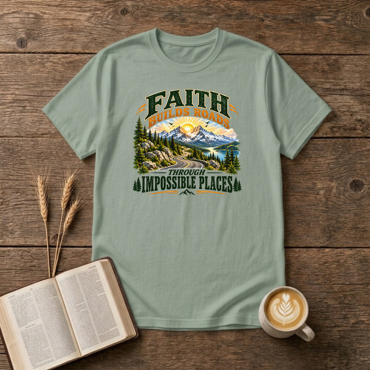 Faith Builds Roads Through Impossible Places design with dark green and orange lettering around a winding mountain road, evergreen forest, lake and sunrise on a Sage unisex T-Shirt