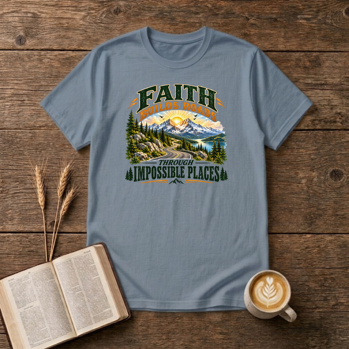 Faith Builds Roads Through Impossible Places design with dark green and orange lettering around a winding mountain road, evergreen forest, lake and sunrise on a Stone Blue unisex T-Shirt