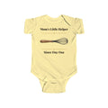 Mom's Little Helper Since Day One baby bodysuit in Butter on a wood table with a knit blanket, wooden rattle and open Bible