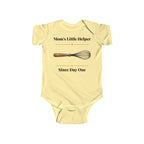 Mom's Little Helper Since Day One baby bodysuit in Butter on a wood table with a knit blanket, wooden rattle and open Bible