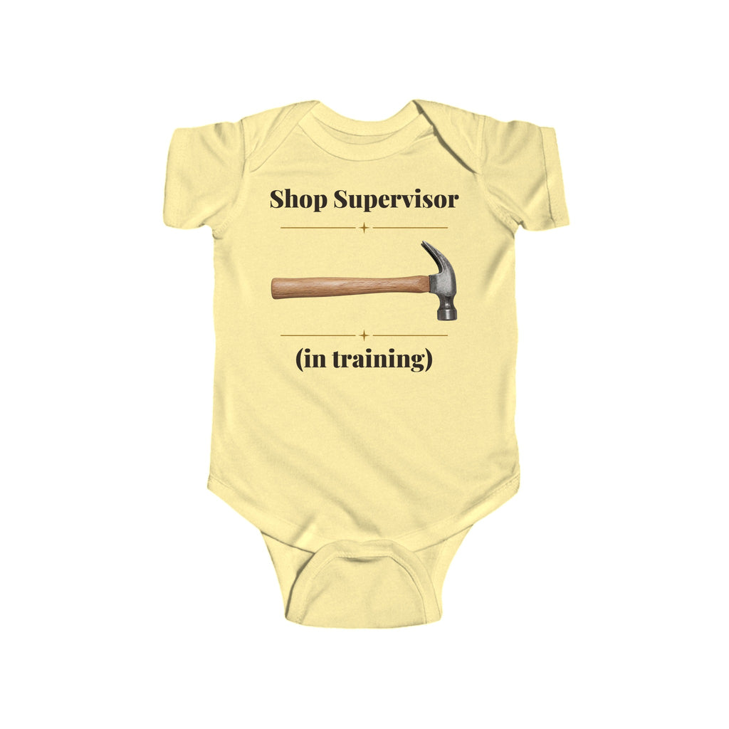 Shop Supervisor in Training baby bodysuit in Butter on a wood table with a knit blanket, wooden rattle and open Bible