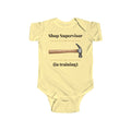 Shop Supervisor in Training baby bodysuit in Butter on a wood table with a knit blanket, wooden rattle and open Bible