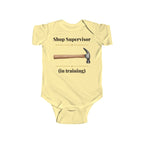 Shop Supervisor in Training baby bodysuit in Butter on a wood table with a knit blanket, wooden rattle and open Bible